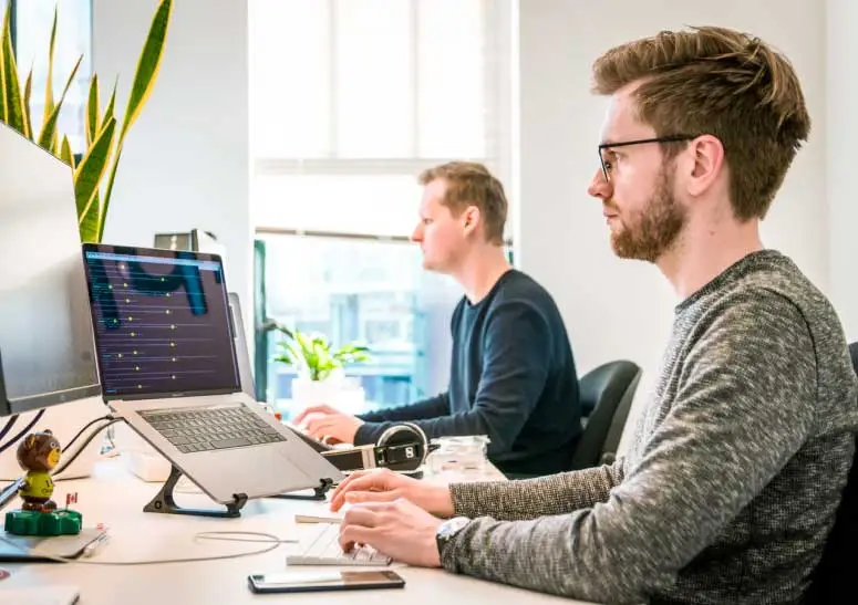 Two men working at desks in an office, both focused on their computers, with one using a laptop on a stand and the other on a desktop setup.